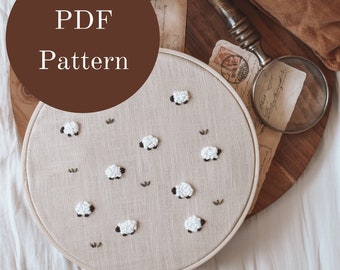 May include: A close-up of a beige embroidery hoop with a white sheep design. The hoop is on a wooden tray with a magnifying glass and vintage postcards. The text "PDF Pattern" is on a brown circle.