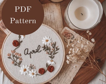 May include: A white embroidery hoop with a floral design and the word "April" stitched in black thread. The hoop is on a wooden tray with a candle, dried flowers, and vintage postcards.
