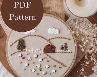May include: A brown wooden embroidery hoop with a beige fabric inside. The fabric has an embroidered scene of a small red house with a white fence, green trees, white clouds, and white sheep in a field. The hoop is on a wooden surface with a white candle and dried flowers.