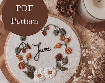 May include: A PDF pattern for an embroidery hoop featuring a floral design with strawberries, daisies, and the word "June" in cursive. The hoop is on a wooden surface with dried flowers and a candle.