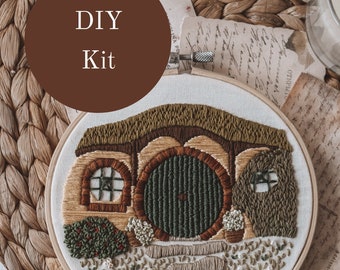 May include: DIY Kit featuring a hand-stitched embroidery hoop with a brown and green hobbit house with a round green door. The hoop is on a woven brown surface.