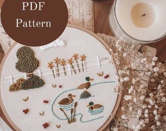 May include: A white embroidery hoop with a hand-stitched design of ducks swimming in a pond with a green tree and a white picket fence. The embroidery is on a beige fabric and the hoop is made of wood.