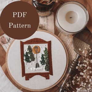 Op de afbeelding: Een witte borduurring met een bruin houten frame, met een kruissteekontwerp van een besneeuwde scène met een lantaarnpaal en bomen. De ring ligt op een houten oppervlak met een kaars, een vergrootglas en gedroogde bloemen.