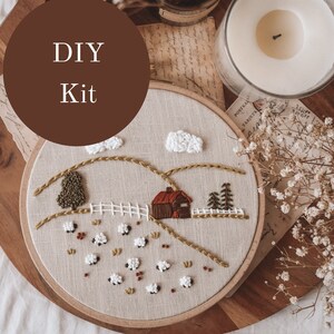 Op de afbeelding: DIY borduurset met een charmante landelijke scène met een klein huis, glooiende heuvels, pluizige wolken en een kudde schapen. De set wordt gepresenteerd op een houten dienblad met een kaars en gedroogde bloemen.