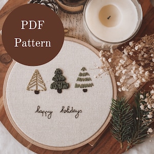 Op de afbeelding: Een borduurring met drie geborduurde kerstbomen en de tekst "happy holidays". De ring ligt op een houten oppervlak, met een kaars, dennenappel en droogbloemen op de achtergrond. De tekst "PDF Pattern" staat in een bruine cirkel.