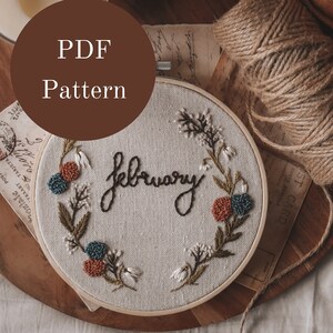 Op de afbeelding: Een PDF-patroon voor een borduurring met het woord "Februari" geborduurd in zwart garen en omringd door een krans van groene bladeren en kleurrijke bloemen.