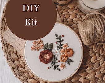 May include: DIY embroidery kit featuring a floral design with orange, red, and white flowers on a white fabric hoop. The hoop is on a woven brown mat.