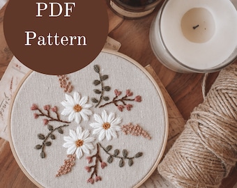 May include: A white embroidery hoop with a floral design featuring white daisies, green leaves, and pink stems. The hoop is on a wooden surface with a candle, a jar, and twine.