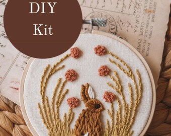 May include: A DIY embroidery kit featuring a brown bunny surrounded by orange flowers and tall, golden grass. The design is set in a wooden hoop. The text "DIY Kit" is in a brown circle at the top.