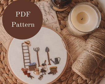 May include: A PDF pattern for a hand-stitched embroidery hoop featuring a ladder, gardening tools, and potted plants. The hoop is on a woven straw surface with a candle, twine, and a vintage postcard.