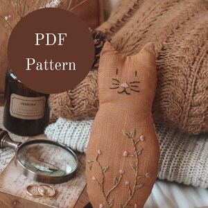 Op de afbeelding: Een PDF-patroon voor een bruin linnen kattenknuffeltje met geborduurde details. Het knuffeltje ligt op een bruin gebreid dekentje met een boek en een vergrootglas.