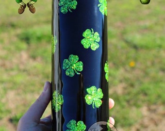 Puede incluir: Un vaso negro decorado con brillantes tréboles verdes. El vaso tiene el texto "St Patrick's day tumbler". También se ve un dibujo de un duende y un arcoíris con un pote de oro.