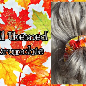 May include: Fall themed scrunchie with a red, orange, and yellow leaf pattern. The scrunchie is tied around a ponytail of gray hair.