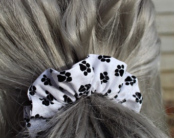 May include: White scrunchie with black paw print design. The scrunchie is tied around a ponytail of gray hair. The scrunchie is labeled "The Carolina Crow"