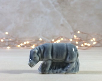 May include: A small, gray ceramic figurine of a rhinoceros. The rhinoceros is depicted in a walking pose with its head lowered.