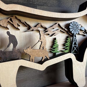 May include: A wooden wall decoration in the shape of a pig with a layered design. The pig is brown and features a silhouette of a person riding a pig, a windmill, and trees.