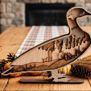 May include: A multi-layered wooden duck silhouette featuring a laser-cut scene of a hunter with a dog in a forest. The design includes cutouts of flying ducks in the neck area and stands on a flat wooden base with a natural wood grain finish.