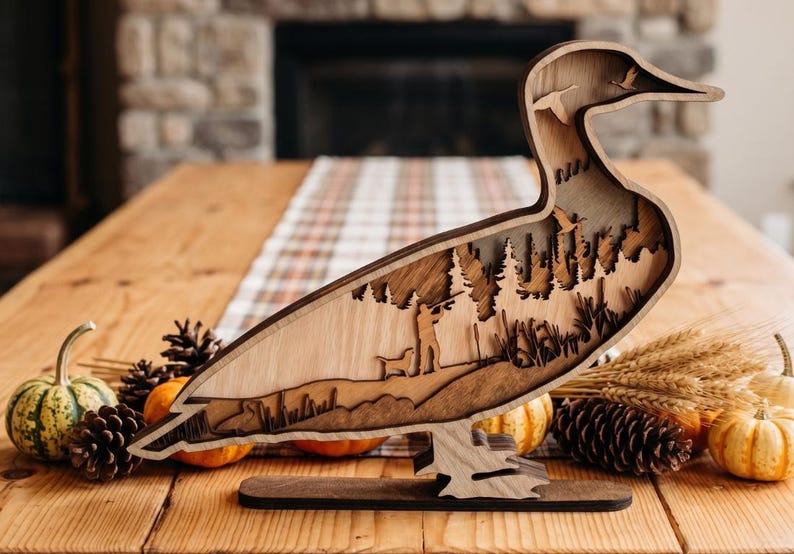 May include: A multi-layered wooden duck silhouette featuring a laser-cut scene of a hunter with a dog in a forest. The design includes cutouts of flying ducks in the neck area and stands on a flat wooden base with a natural wood grain finish.