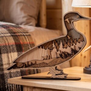 May include: A multi-layered wooden sculpture shaped like a duck, featuring a laser-cut scene of a hunter with a dog in a forest, with additional silhouettes of flying birds integrated into the upper body and a flat base for stability.