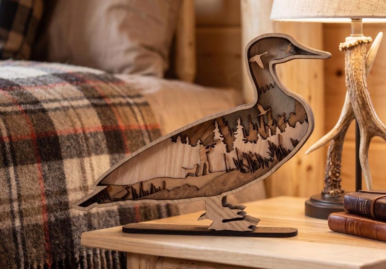 May include: A multi-layered wooden sculpture shaped like a duck, featuring a laser-cut scene of a hunter with a dog in a forest, with additional silhouettes of flying birds integrated into the upper body and a flat base for stability.