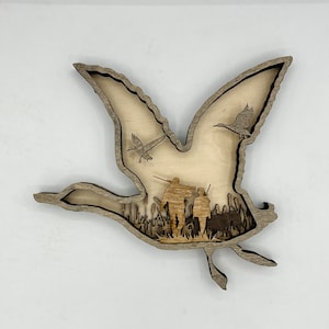 May include: A wooden cutout of a flying goose with two smaller geese flying above it. The cutout is layered to show a scene of hunters in a field of tall grass.