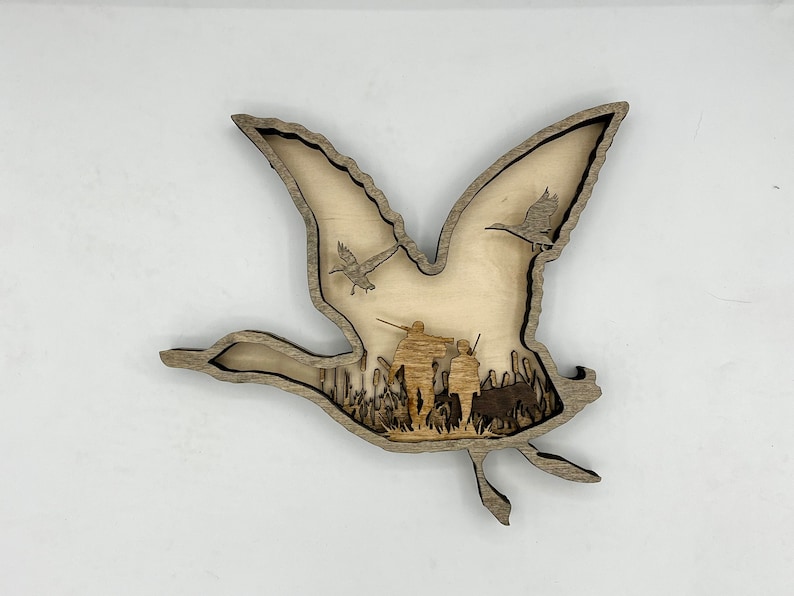 May include: A wooden cutout of a flying goose with two smaller geese flying above it. The cutout is layered to show a scene of hunters in a field of tall grass.