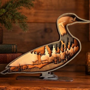 May include: A multi-layered wooden duck silhouette featuring a laser-cut scene of a hunter with two dogs in a forest. The design includes intricate cutouts of pine trees, tall grasses, and a flying bird, all finished in warm brown wood tones.