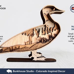 May include: A multi-layered wooden duck silhouette featuring a laser-cut landscape scene with a person and dog. Text includes 6 LAYERS OF PREMIUM WOOD, MADE IN COLORADO, UNIQUE WOOD CHARACTER, NATURAL WOOD IMPERFECTIONS, and Bunkhouse Studio - Colorado Inspired Decor.