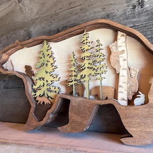 K&ouml;nnte beinhalten: Eine h&ouml;lzerne B&auml;ren-Silhouetten-Wanddekoration mit einem geschichteten Design. Der B&auml;r ist braun und hat einen Ausschnitt einer Waldszene im Inneren. Die Szene umfasst B&auml;ume, B&auml;ren und ein Kaninchen.