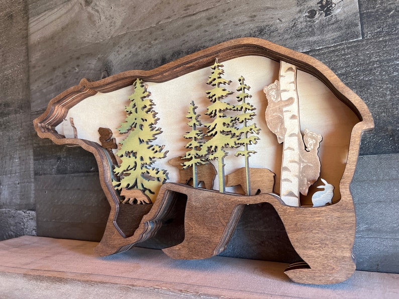 K&ouml;nnte beinhalten: Eine h&ouml;lzerne B&auml;ren-Silhouetten-Wanddekoration mit einem geschichteten Design. Der B&auml;r ist braun und hat einen Ausschnitt einer Waldszene im Inneren. Die Szene umfasst B&auml;ume, B&auml;ren und ein Kaninchen.