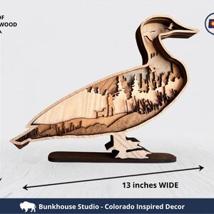 May include: A layered wooden duck sculpture featuring 7 layers of premium wood, measuring 10 inches tall and 13 inches wide. The interior cutout depicts a forest scene with trees and a person. Text includes 7 LAYERS OF PREMIUM WOOD, MADE IN COLORADO, and Bunkhouse Studio - Colorado Inspired Decor.