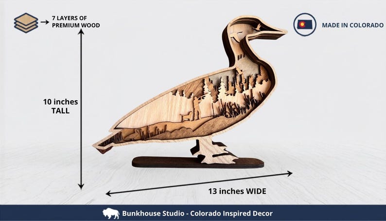 May include: A layered wooden duck sculpture featuring 7 layers of premium wood, measuring 10 inches tall and 13 inches wide. The interior cutout depicts a forest scene with trees and a person. Text includes 7 LAYERS OF PREMIUM WOOD, MADE IN COLORADO, and Bunkhouse Studio - Colorado Inspired Decor.