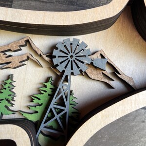 May include: A wooden layered wall art piece featuring a black windmill, green trees, and brown mountains. The piece is crafted from multiple layers of wood, creating a 3D effect.