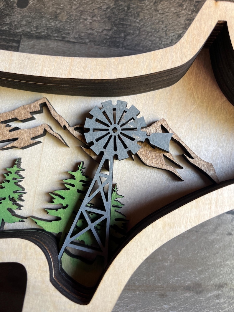 May include: A wooden layered wall art piece featuring a black windmill, green trees, and brown mountains. The piece is crafted from multiple layers of wood, creating a 3D effect.