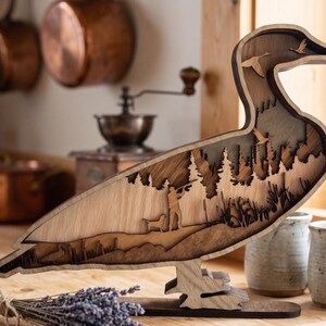 May include: A layered wooden duck-shaped sculpture featuring a cutout scene of a hunter with a dog in a forest. The design includes silhouettes of flying ducks, tall pine trees, and reeds, crafted from light and dark wood tones with a smooth finish.