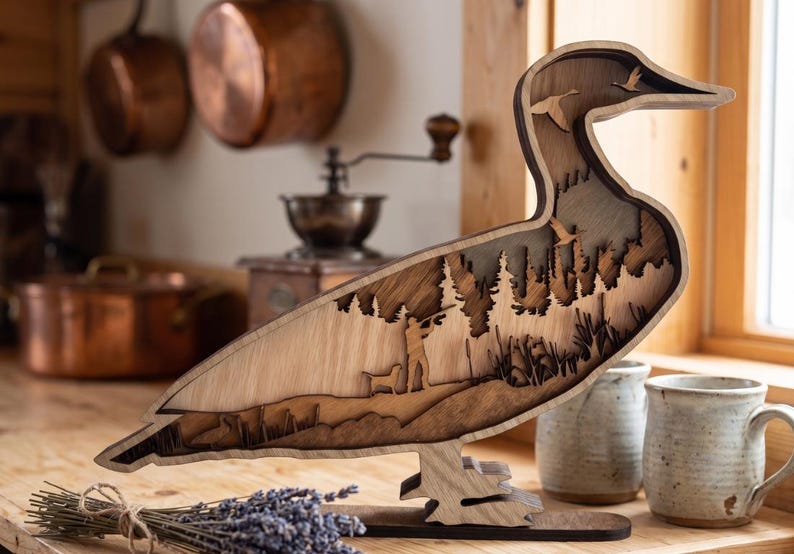 May include: A layered wooden duck-shaped sculpture featuring a cutout scene of a hunter with a dog in a forest. The design includes silhouettes of flying ducks, tall pine trees, and reeds, crafted from light and dark wood tones with a smooth finish.