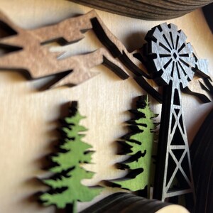 May include: A wooden wall art piece with a 3D design of a windmill, trees, and mountains. The windmill is black and the trees are green. The mountains are brown.