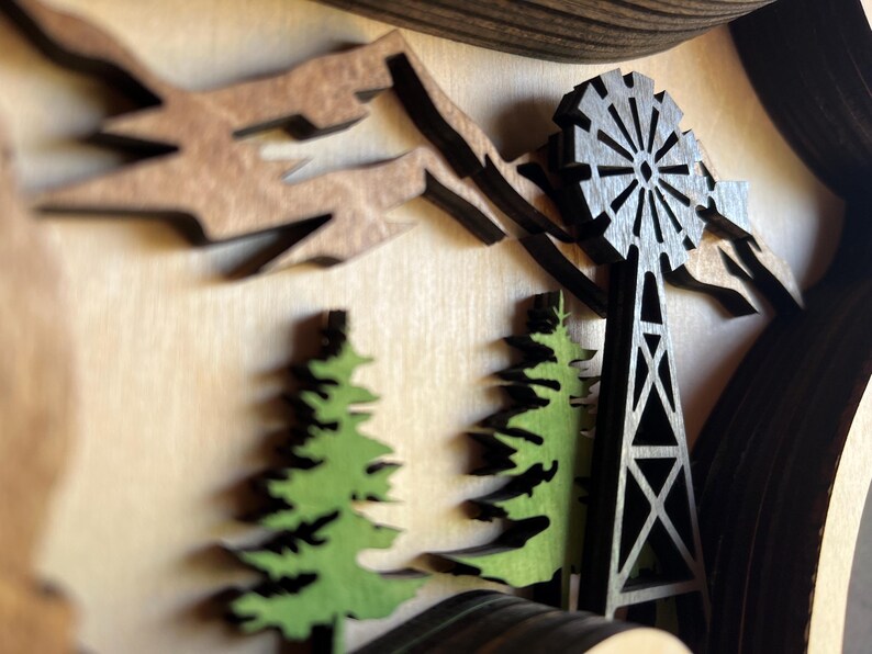 May include: A wooden wall art piece with a 3D design of a windmill, trees, and mountains. The windmill is black and the trees are green. The mountains are brown.