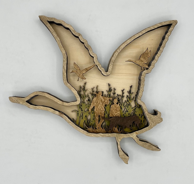 May include: A wooden cutout of a flying goose with a scene of hunters and a dog in the middle. The hunters are walking through tall grass and the dog is running beside them.