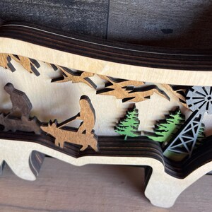 May include: A wooden layered art piece depicting a scene with two figures riding animals, trees, and a windmill. The piece is made of multiple layers of wood, creating a 3D effect.