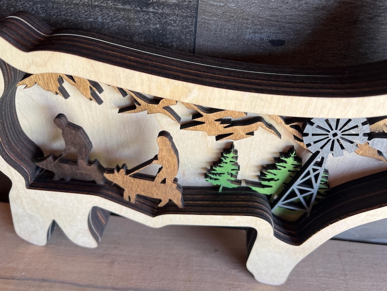 May include: A wooden layered art piece depicting a scene with two figures riding animals, trees, and a windmill. The piece is made of multiple layers of wood, creating a 3D effect.