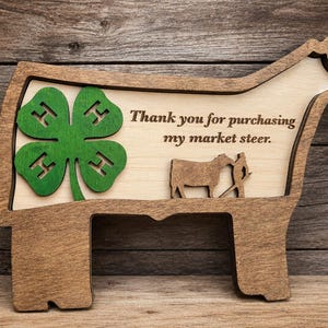 May include: A wooden sign shaped like a steer featuring a green four-leaf clover with an H on each leaf. The sign includes the engraved text, Thank you for purchasing my market steer, and a small cutout of a person leading a steer.
