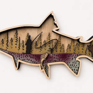 May include: A multi-layered wooden wall art piece shaped like a fish. The interior features a laser-cut scene with a silhouette of a person fishing, a forest of pine trees, a leaping fish, and a moose, all set against a light wood background.