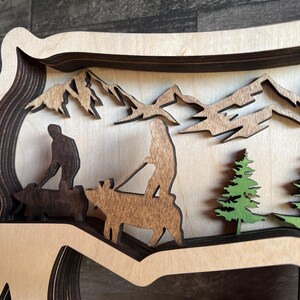 May include: A wooden layered art piece depicting a mountain scene with two figures riding on animals. The figures are silhouetted in brown, and the animals are brown and white. The mountains are brown and the trees are green.