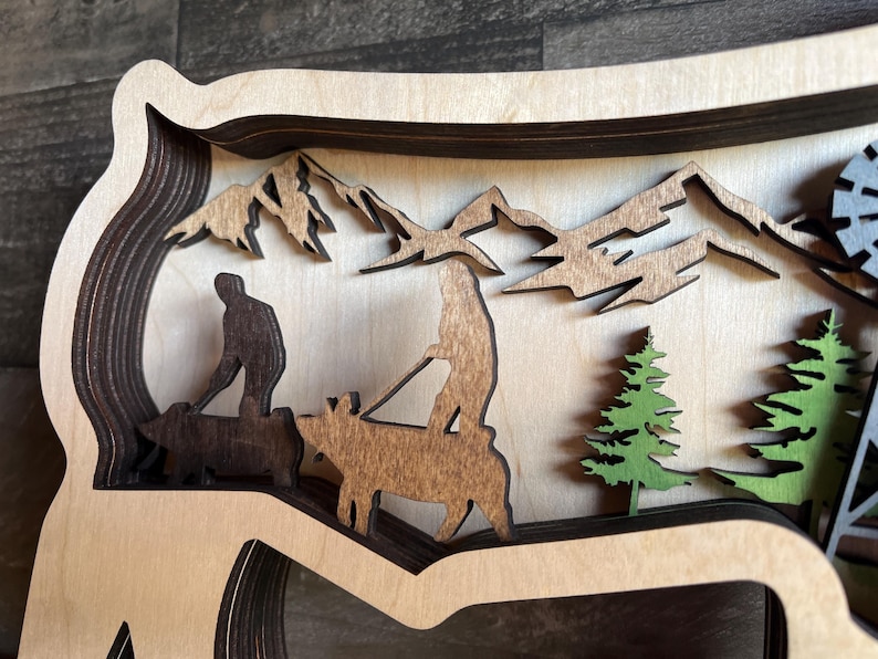 May include: A wooden layered art piece depicting a mountain scene with two figures riding on animals. The figures are silhouetted in brown, and the animals are brown and white. The mountains are brown and the trees are green.