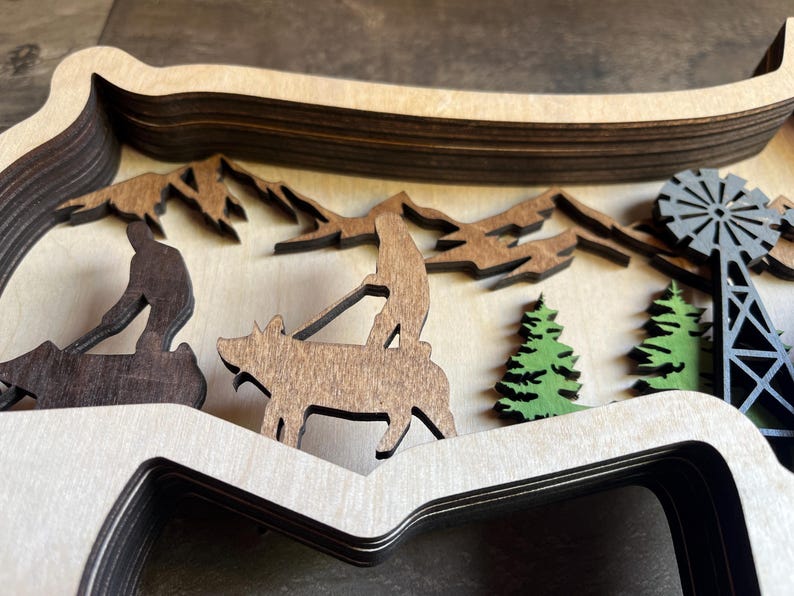 May include: A layered wooden wall art piece depicting a rural scene with a pig, two figures, trees, and a windmill. The piece is made of multiple layers of wood, creating a 3D effect.