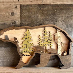 K&ouml;nnte beinhalten: Eine h&ouml;lzerne B&auml;ren-Silhouetten-Wanddekoration mit einem geschichteten Design. Der B&auml;r ist braun und hat einen Ausschnitt einer Waldszene mit B&auml;umen, B&auml;ren und anderen Waldtieren.