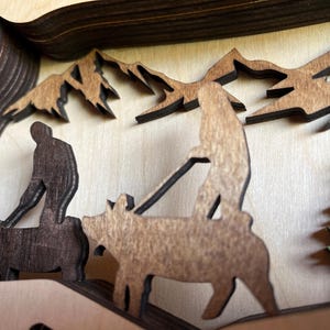 May include: A wooden layered art piece depicting a family of three walking through a forest. The family is made up of two adults and a child. The adults are walking side by side, while the child is walking behind them. The forest is made up of trees and mountains. The art piece is painted in shades of brown and green.