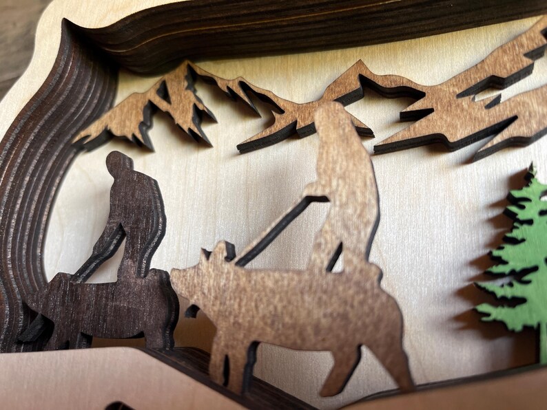 May include: A wooden layered art piece depicting a family of three walking through a forest. The family is made up of two adults and a child. The adults are walking side by side, while the child is walking behind them. The forest is made up of trees and mountains. The art piece is painted in shades of brown and green.