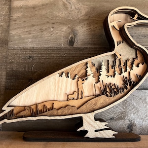 May include: A multi-layered wooden wall art piece shaped like a duck, featuring a laser-cut scene of a hunter with a dog in a forest. The design includes silhouettes of flying birds and reeds, with varying shades of natural wood grain and dark brown edges.